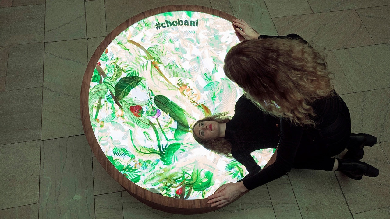 Young woman looking into the final Chobani selfie mirror, with colorful and lush plant illustrations in the background