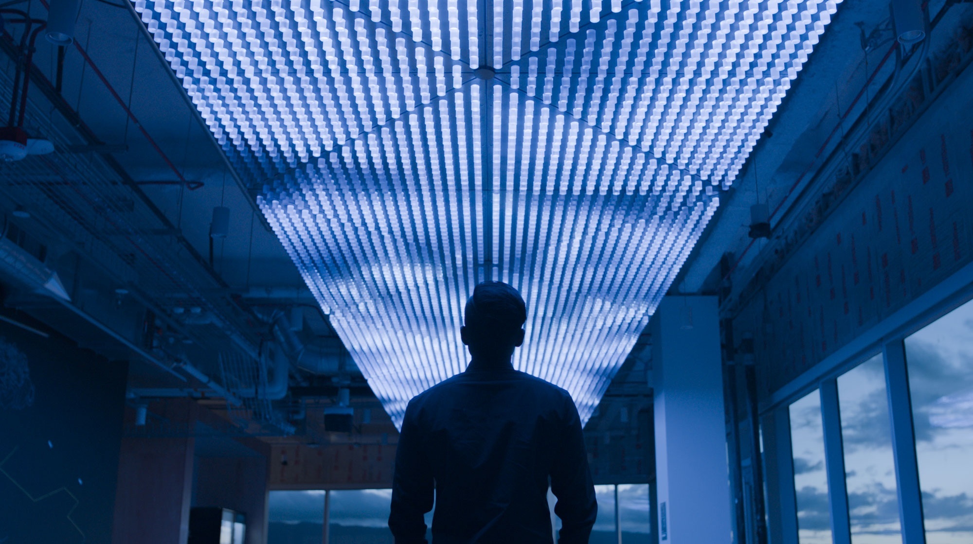 Symmetrical image of silhouetted man looking up at ceiling installation made of repeating protruding light modules that simulate waves and create visualisation