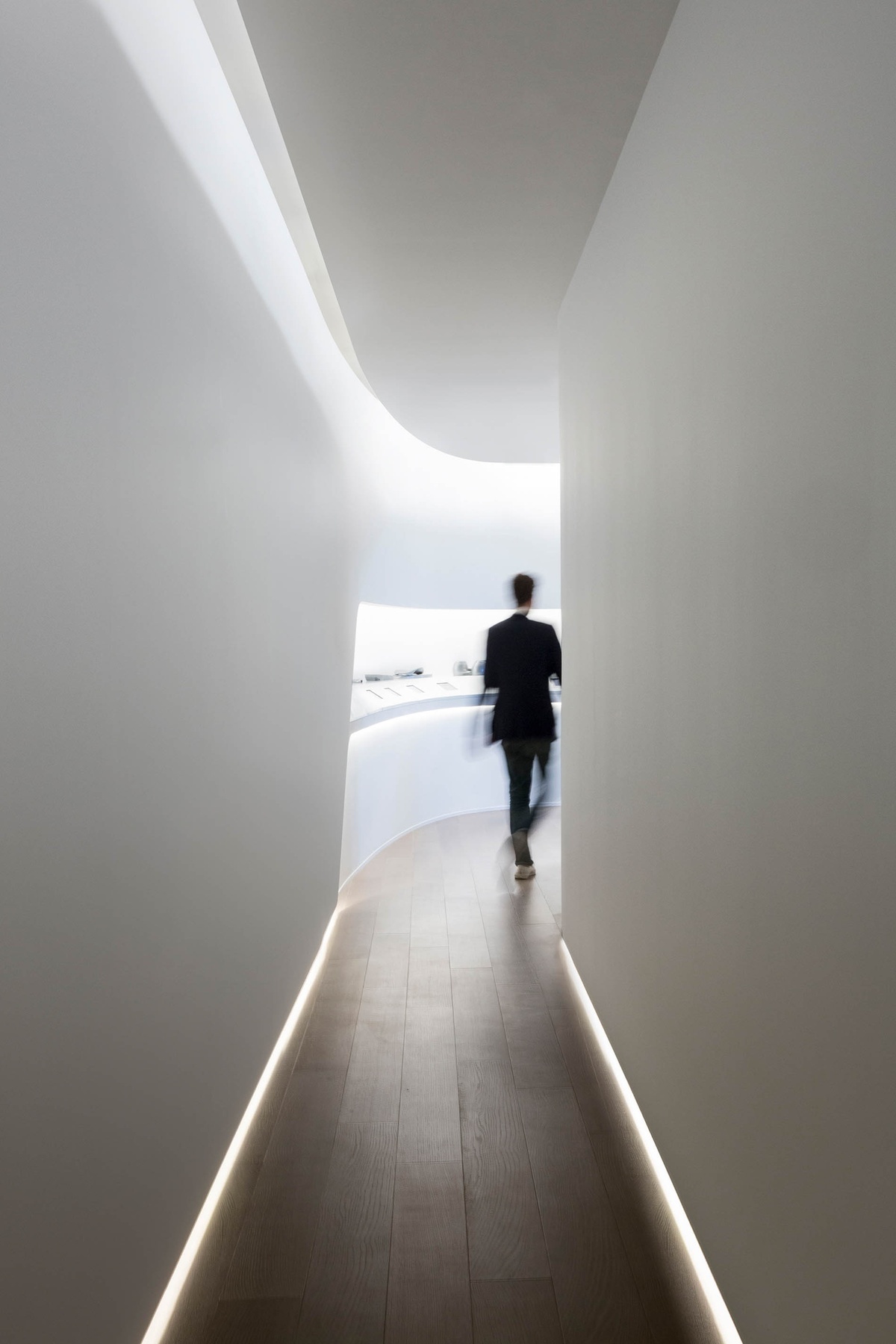 Image of person walking to the end of a long hallway, which opens up to a well-lit space