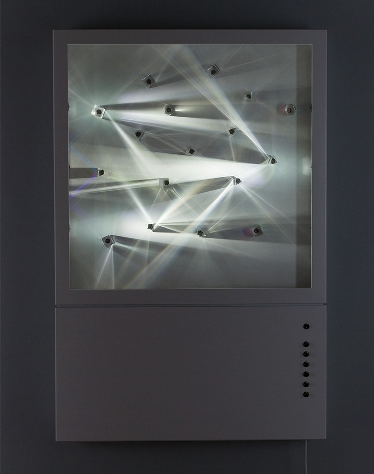 A rectangular device containing a blank "canvas", transparent prisms and one light that emits from several points in the frame, creating interesting visual pattern of light interaction