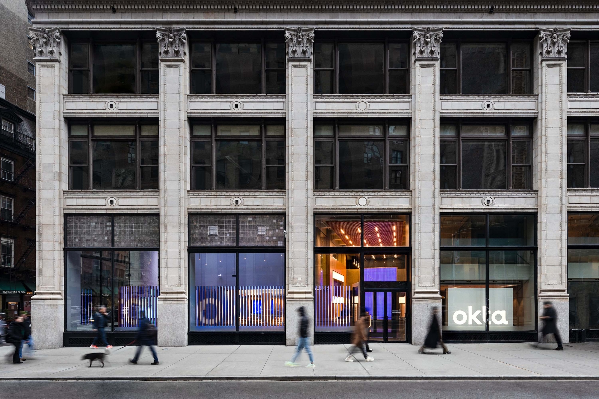 Image of classical architectural exterior of the building that houses the Okta sales center with a number of pedestrians walking past, and the interactive Okta animation visible from the window