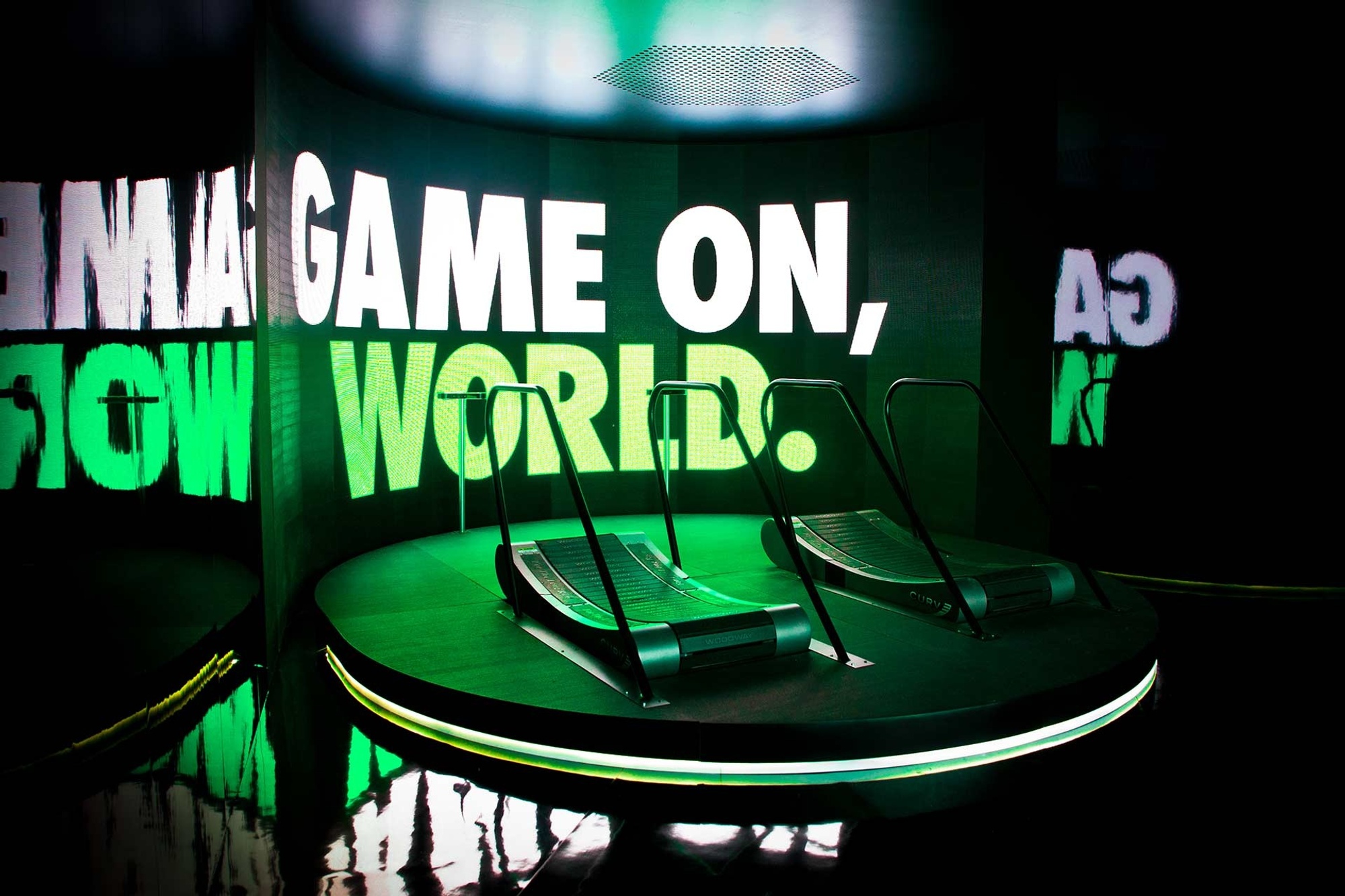 Two treadmills on a raised platform against a screen that says "Game on, world" and reflects neon green light onto the platform
