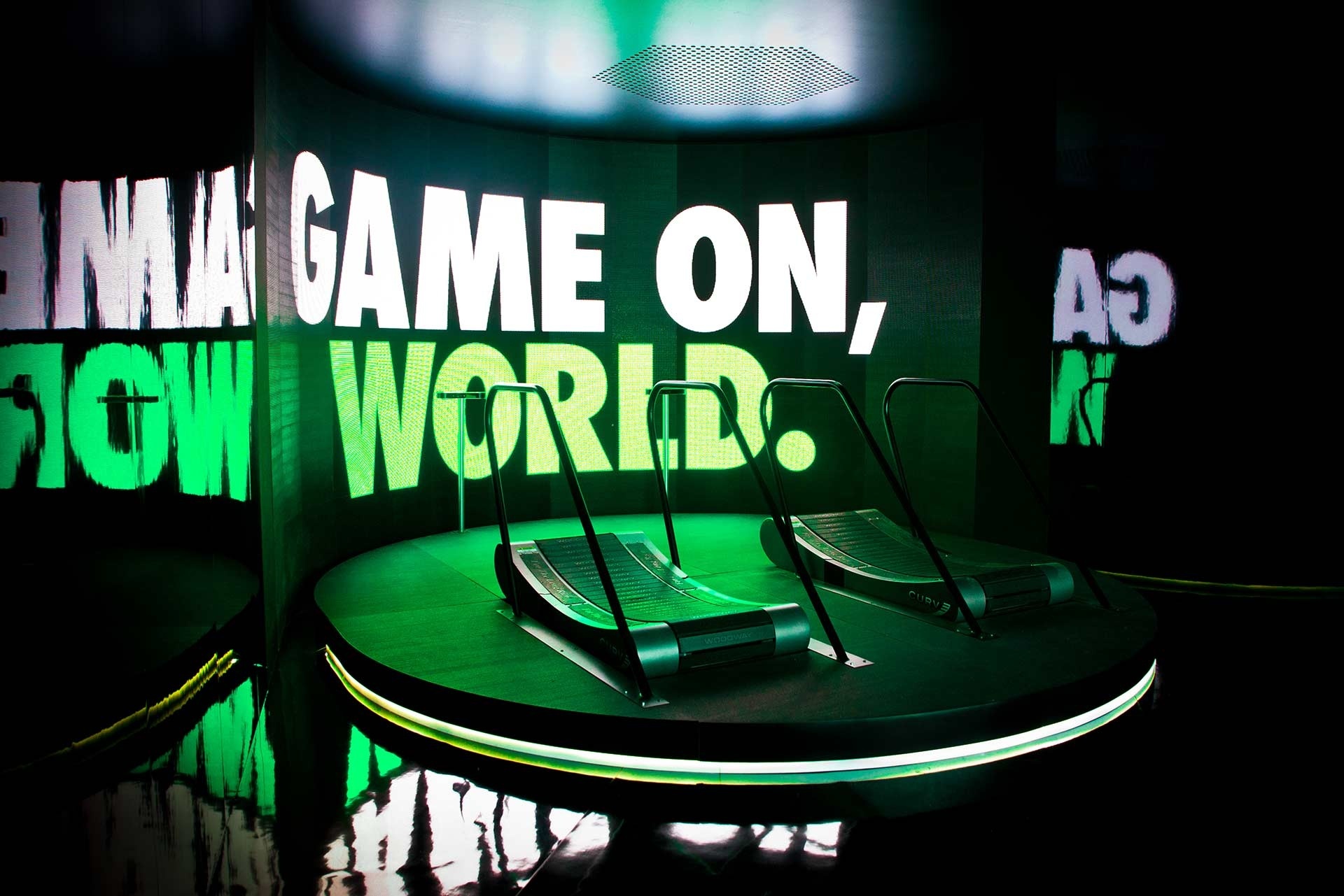 Two treadmills on a raised platform against a screen that says "Game on, world" and reflects neon green light onto the platform