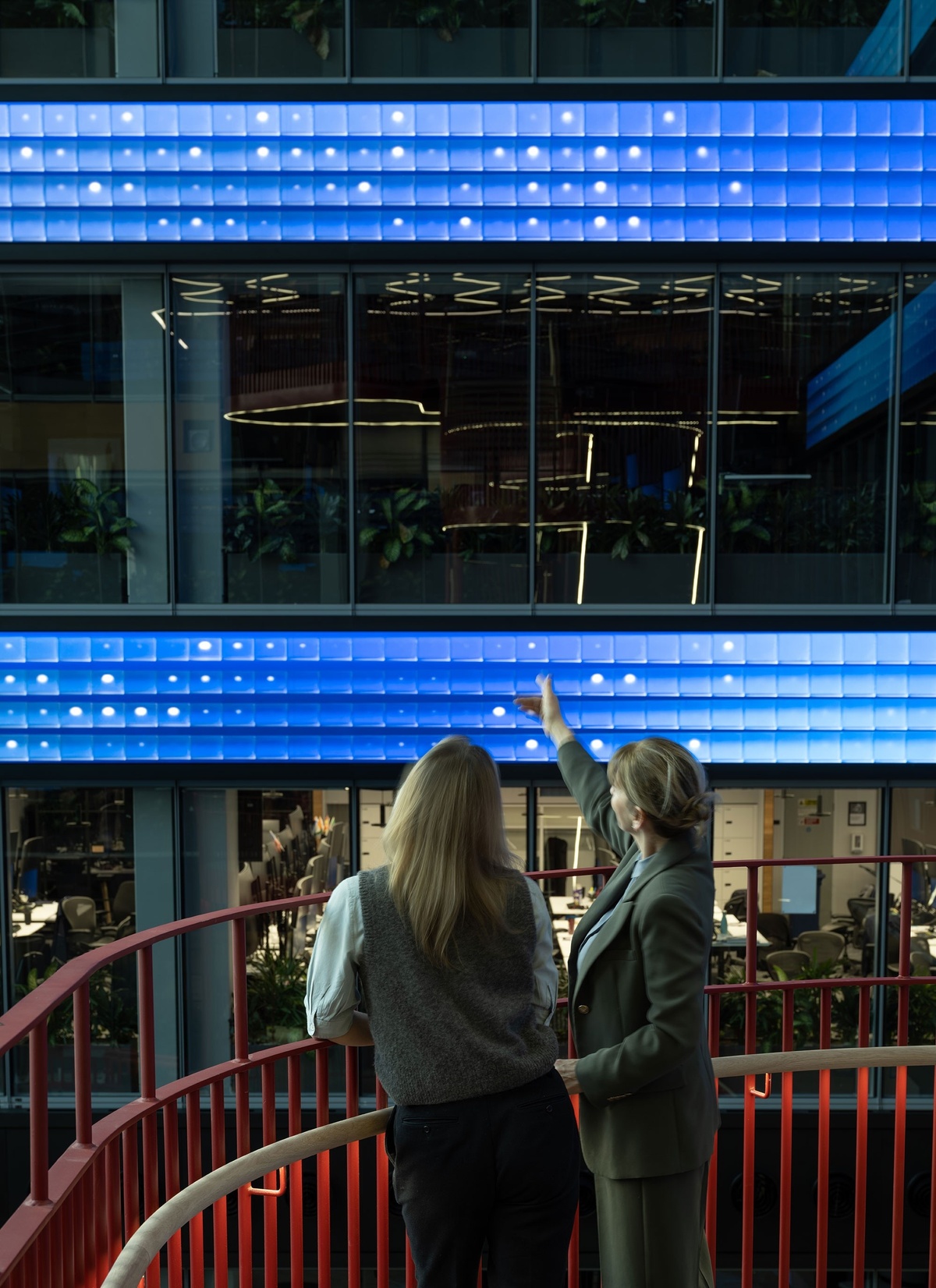 Two women on the stairwell, leaning against the banister and looking out at the installation in the atrium